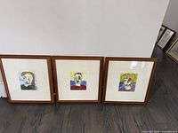 Three framed abstract portraits of women with colorful and whimsical styles, sitting on wood floor against white wall.