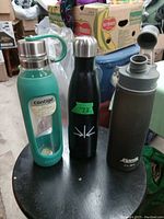 Three reusable water bottles on a black stool: Contigo glass bottle in teal, black stainless steel bottle with '73' sticker, gray CamelBak plastic bottle