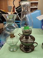 Photo showing two vintage oil lamps: one clear glass with brass burner and flared chimney, one dark metal base with handle and tall chimney.