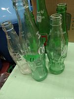Four vintage glass soda bottles: two clear Coca-Cola bottles, one green 7 Up bottle, and one additional green glass bottle