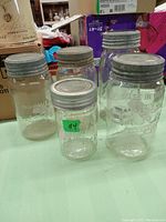 Photo showing four vintage glass jars of varying sizes with metal lids placed on green surface. Background includes cardboard boxes and other items.