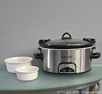 Full view of oval electric Crock-Pot with stainless steel body, glass lid, and black handles alongside two white round Corningware dishes of different sizes on a green glass table.