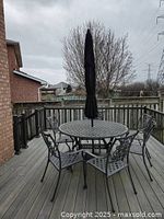 Full view of round cast metal patio dining table with four matching chairs and black umbrella closed in center of table on outdoor deck.