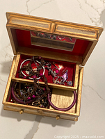 Open wooden jewellery box showing red velvet lining with assortment of costume jewelry including bracelets and earrings in upper and lower compartments.