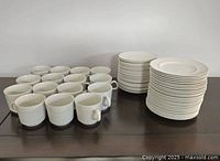 Shows a group of white Johnson Brothers Athena cups arranged in front with two stacks of saucers and side plates in the back on a wooden surface.