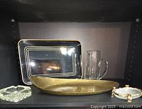 Five vintage items displayed on a shelf: a clear faceted crystal ashtray, a black rectangular tray with gold trim, a hammered brass dish, a clear glass pitcher with handle, and a small round trinket dish with gold gilt bamboo detail.