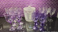 Multiple purple and clear hard plastic outdoor glasses arranged on a countertop with purple tile backsplash. Includes goblet and wine glass shapes.