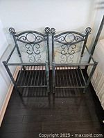 Pair of wrought iron nightstands with clear glass tops, verdigris finish, and decorative scrollwork against a wall by a bed.