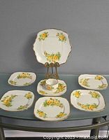 Set displayed on table including six luncheon plates, one cake plate on a stand, one open sugar bowl, and one cup. All pieces decorated with yellow rose floral designs and gilt edges.