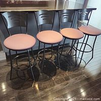 Four stools with black metal frames, wavy backrests, and light brown upholstered seats shown side by side on a wood floor
