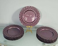 Full view of twelve purple glass chargers with textured basketweave pattern arranged as two neat stacks on a glass surface and one plate displayed upright on a stand.