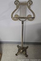 Full view of the vintage brass music stand showing lyre-shaped music holder, adjustable pole, and ornate cast base on a tiled floor.