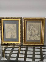 Two framed pencil sketches displayed side by side on a glass tabletop; one depicts hands, the other a woman in period clothing.