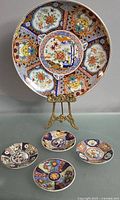 One large Imari platter displayed on a decorative stand behind four smaller Imari plates laid out flat showing the detailed colorful patterns.