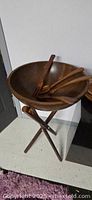 Large wooden standing salad bowl on tripod base with wooden salad servers inside bowl.