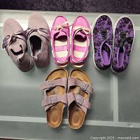 Top view of four pairs of women's casual shoes on a glass surface: lilac suede Birkenstock sandals, fuchsia Crocs loafers, orchid Skechers mesh shoes, and royal purple lace print canvas sneakers.