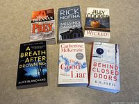 Six paperback thriller mystery novels displayed spread in two rows on a carpeted floor. Covers visible showing titles and authors.