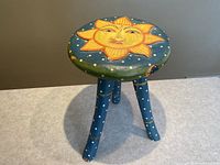 Full view of a small wooden stool with a sun motif on the seat surface, blue legs decorated with celestial dots and crescents.