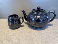 Photo of brown glazed redware creamer and tea pot with handles, featuring silver overlay floral designs on both pieces, placed on a counter.