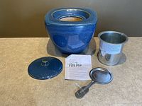 Blue Green Earth ceramic fire pot with lid, metal insert, pot snuffer, and manual laid out on table surface.