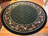 Full view of the round area rug on wooden floor with dark blue border, green lattice pattern center, and fruit and flower detail band.