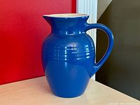 Blue ceramic pitcher with tan interior on a light wood surface against a red wall.