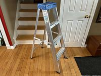 Side view of aluminum ladder showing three steps, blue top tray, rubber feet, and cross braces.