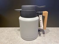 Front angle photo showing taupe insulated coffee carafe with wooden handle and dark grey lid with lever for pouring, original tag visible.