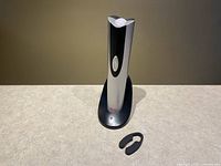 Oster electric wine cork bottle opener on charging base with foil cutter ring on the side, shown powered off on a tabletop against a brown background.