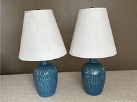 Pair of blue ceramic table lamps with beige shades shown side by side on a beige surface against a gray background, lamps off.