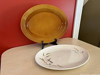 White oval platter with wheat design on a black display stand in front of an ochre brown oval platter against red wall