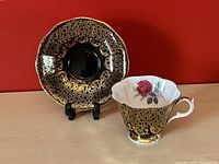 Tea cup and matching saucer displaying black and gold intricate design, pink rose detail inside the cup, on wooden surface