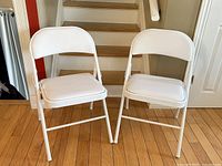 Front view of two white folding chairs placed on hardwood floor near staircase, showing padded seat and backrest.