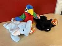 Photo showing three Ty Beanie Babies - Velvet Panther (black), Peanut Elephant (light gray with pink ears), Jabber Parrot (multicolored) with original TY heart tags attached.