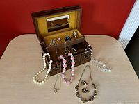 Vintage wooden music jewelry box open showing velvet lined interior, mirror, and various necklaces and brooches displayed inside and hanging outside the box.
