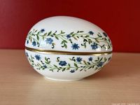 Egg-shaped porcelain trinket box with blue floral and vine decoration, shown closed from an angled side view with red background.