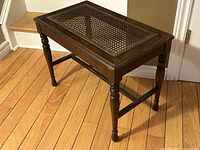 Side angled view of vintage walnut wood bench with cane top on hardwood floor