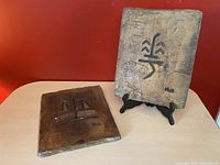 Two rectangular concrete stone plaques displayed on a table against a red wall. The beige plaque with Zen flower symbol is on a black stand, and the dark brown plaque with Zen meditation symbol lies flat.