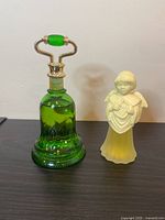 Front view of the green glass bell-shaped bottle with silver handle and green bead, next to the yellow frosted glass angel figurine holding a lyre.