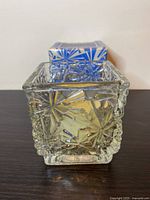 Front view of clear glass square candlette holder with starburst pattern and original box behind.