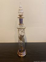 Front view of tall, slender clear glass bottle with gold and blue decorative patterns and stopper.