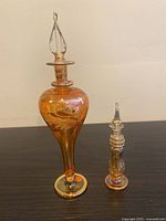 Two iridescent amber glass bottles with detailed gold painted birds and foliage, tall twisted stopper and smaller ornamental stopper, shown together on dark surface