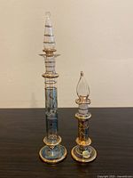 Pair of tall slender Egyptian hand blown glass bottles with elaborate stoppers. Gold banding and colored glass designs visible. Shown on dark surface against neutral wall.