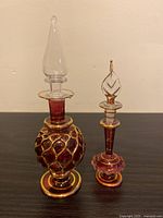 Photo showing two Egyptian hand blown glass bottles with stoppers in red glass with gold accents, positioned side by side on a dark surface against a white background.