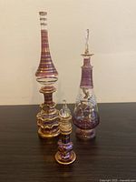 Three purple Egyptian hand blown glass bottles with stoppers arranged side by side on a flat surface against a neutral wall, showing differing heights and stopper designs