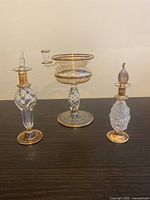 Three hand blown glass art pieces including two bottles with stoppers and a footed oil burner bowl placed on a dark surface in front of a light wall.