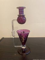 Front angle photo showing the deep purple glass oil burner with gold detailing and clear twisted handle, placed upright against a beige background.