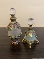 Photo of two frosted glass bottles with ornate jeweled brass overlays and clear faceted stoppers on a dark surface against a plain wall.