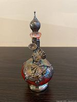 Front view of small red glass bottle with metal stopper and floral silver accents showing detailed metal work on the bulbous body and stopper.