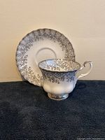 Cup and saucer set displayed on dark cloth with saucer propped behind cup, showing silver chintz leaf pattern and scalloped edges